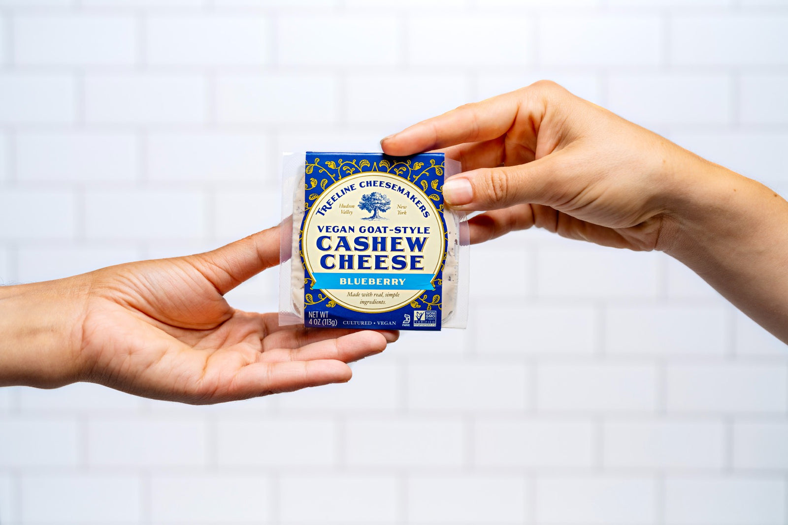 Hands passing a package of Treeline vegan blueberry goat style cheese